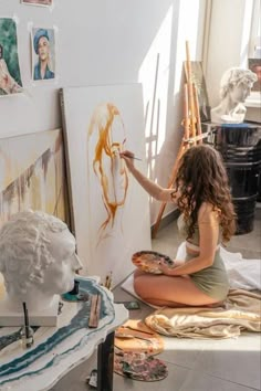 a woman sitting on the floor in front of some paintings and holding a paintbrush