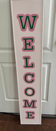 a welcome sign hanging from the side of a white door with pink and green lettering