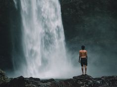 a man standing at the base of a waterfall looking out into the water from below