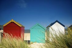 three colorful beach huts sitting on top of a sandy beach next to tall green grass