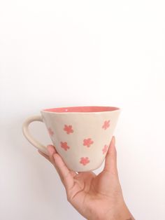 a hand holding a pink and white coffee cup with red flowers on the rim, against a white background