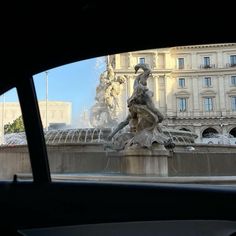 a fountain in the middle of a city with buildings and cars passing by behind it