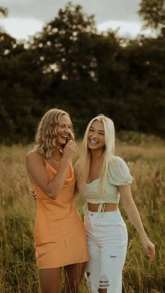 two women standing next to each other in a field with tall grass and trees behind them