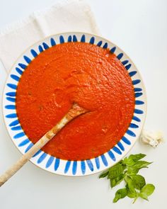 a bowl of tomato soup with a wooden spoon in it and basil on the side