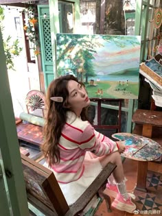 a woman sitting on top of a wooden bench in front of a easel filled with paintings