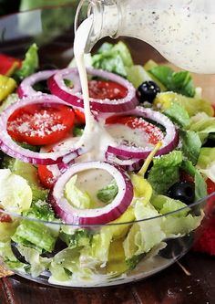 a salad with dressing being poured onto it and topped with lettuce, tomatoes, onions, black olives, cucumbers, red onion and cheese