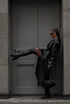 a woman in black boots is leaning against a door with her leg up and holding onto a purse