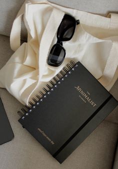 an open notebook and sunglasses are sitting on a couch next to a white bag with black writing