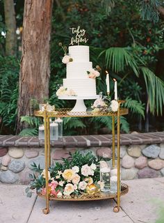 a wedding cake sitting on top of a table next to flowers and greenery in front of a tree