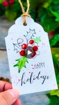 a hand holding a christmas gift tag with holly berries on it and the words happy holidays written in black ink