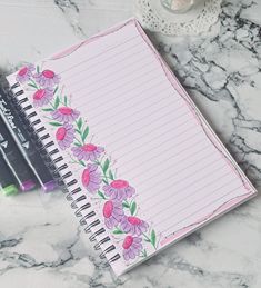 a notepad with pink flowers on it next to two pens and a cup of coffee