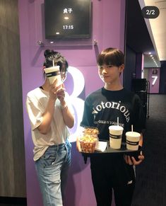 two people standing in front of a purple wall holding cups and plates with food on them