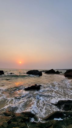 the sun is setting over the ocean with rocks in the foreground and water splashing on the shore