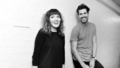 two people standing next to each other in front of a white brick wall and smiling at the camera