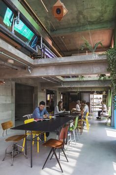 people are sitting at tables in a large room with concrete walls and green plants hanging from the ceiling