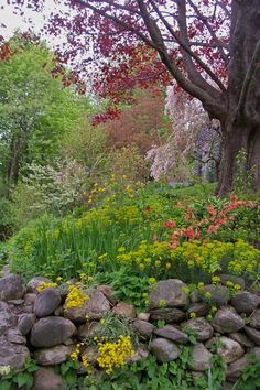 a stone wall in the middle of a garden with flowers growing on it and trees