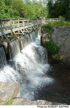 a wooden bridge over a small waterfall with water running down it's sides and people standing on the other side