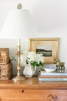 a white vase with flowers on top of a wooden dresser next to a lamp and books