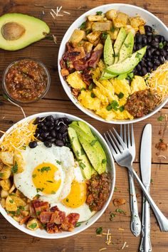 two white bowls filled with breakfast food on top of a wooden table next to an avocado