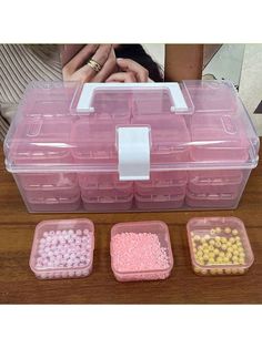 three plastic containers filled with beads on top of a wooden table next to a woman's hand