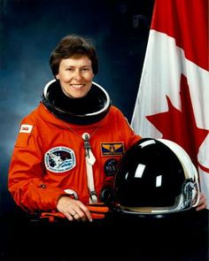 a woman in an orange space suit standing next to a canadian flag and holding a helmet