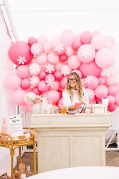 a woman standing behind a counter with pink balloons on the wall and decorations around her