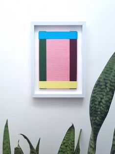 an abstract painting in a white frame hangs on the wall next to a green plant