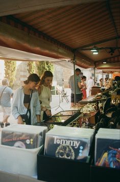 two women are looking at cds on display in a market area with people standing around