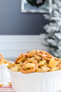 two white bowls filled with cereal and pretzels next to a christmas tree in the background