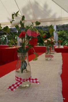 red and white flowers are in glass vases on a long table with gingham cloth