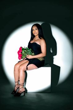 a woman in a black dress sitting on a white block holding flowers and posing for the camera