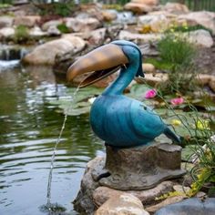 a statue of a bird drinking water from a pond with rocks and flowers around it