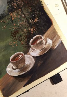 two coffee cups sitting on top of a wooden table next to a painting with trees in the background