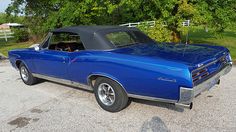 an old blue car parked in a parking lot next to a tree and fenced in area