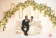 a bride and groom sitting on a couch in front of an arch of greenery