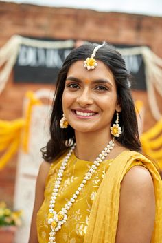 a woman in a yellow sari and pearls necklace smiles at the camera while standing outside