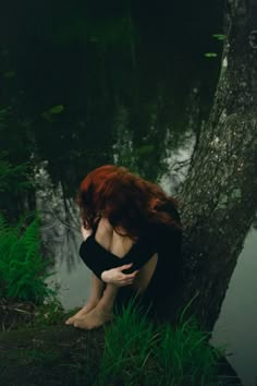 a woman with red hair sitting on the ground next to a tree in front of a body of water