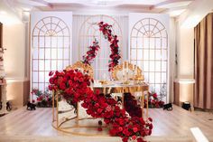 a table with red flowers on it in front of two arched windows and gold chairs