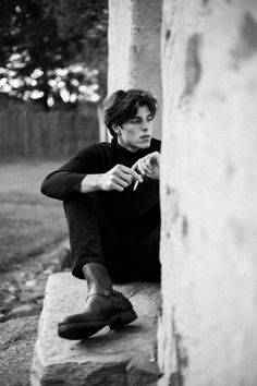 a young man sitting on top of a stone wall next to a wooden fence with his hand under his chin