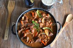 a pot filled with meat and carrots next to a glass of beer on top of a wooden table