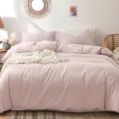 a bed with pink comforter and pillows in a white room next to a wicker chair