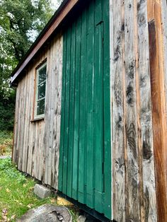 an old wooden shed with green door and windows on the side, in front of some trees