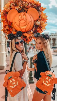 two women standing next to each other with pumpkin backpacks on their shoulders and smiling at the camera