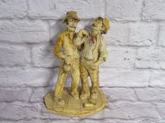 two figurines standing next to each other on a white brick wall with bricks in the background