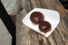 two chocolate covered cupcakes sitting on top of a white paper container next to a wooden table