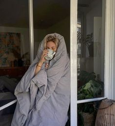 a woman wrapped up in a blanket drinking from a cup while sitting on the porch