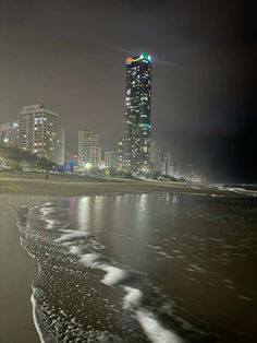 the city is lit up at night by the beach and waves in the foreground