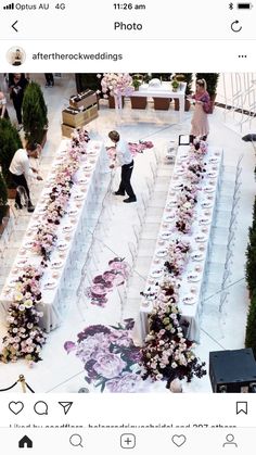 two people standing at the end of a long table covered in flowers and greenery