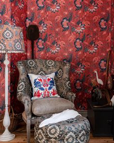 a chair with a pillow on it in front of a red floral wallpapered room