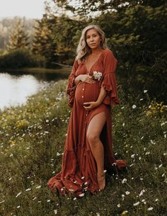 a pregnant woman in a red dress poses for a photo by a lake with wildflowers